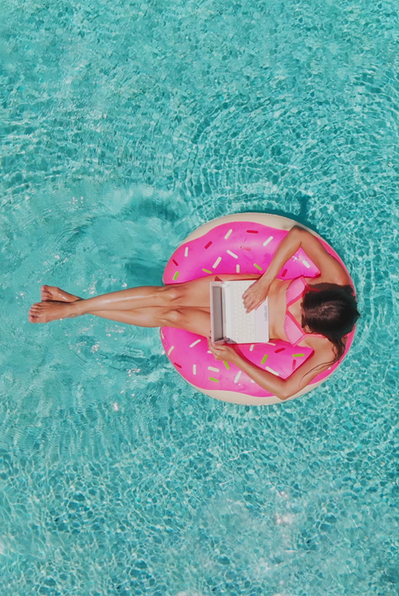 Vue aérienne d'une jeune femme brune nageant sur un gros beignet gonflable avec un ordinateur portable dans une piscine turquoise transparente.