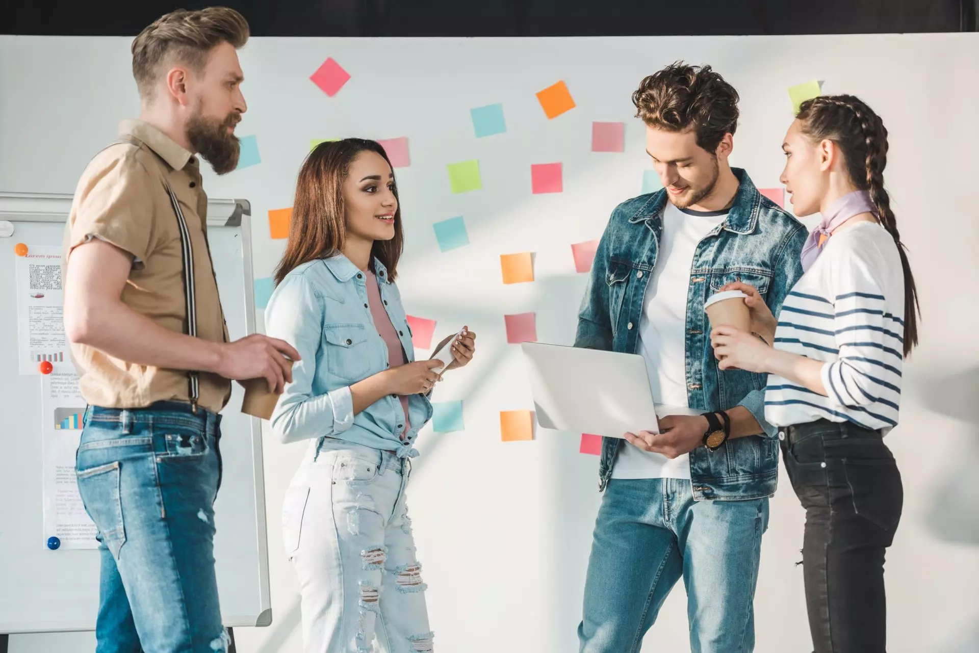 Diverse business team with laptop by white board with sticky notes in light workspace