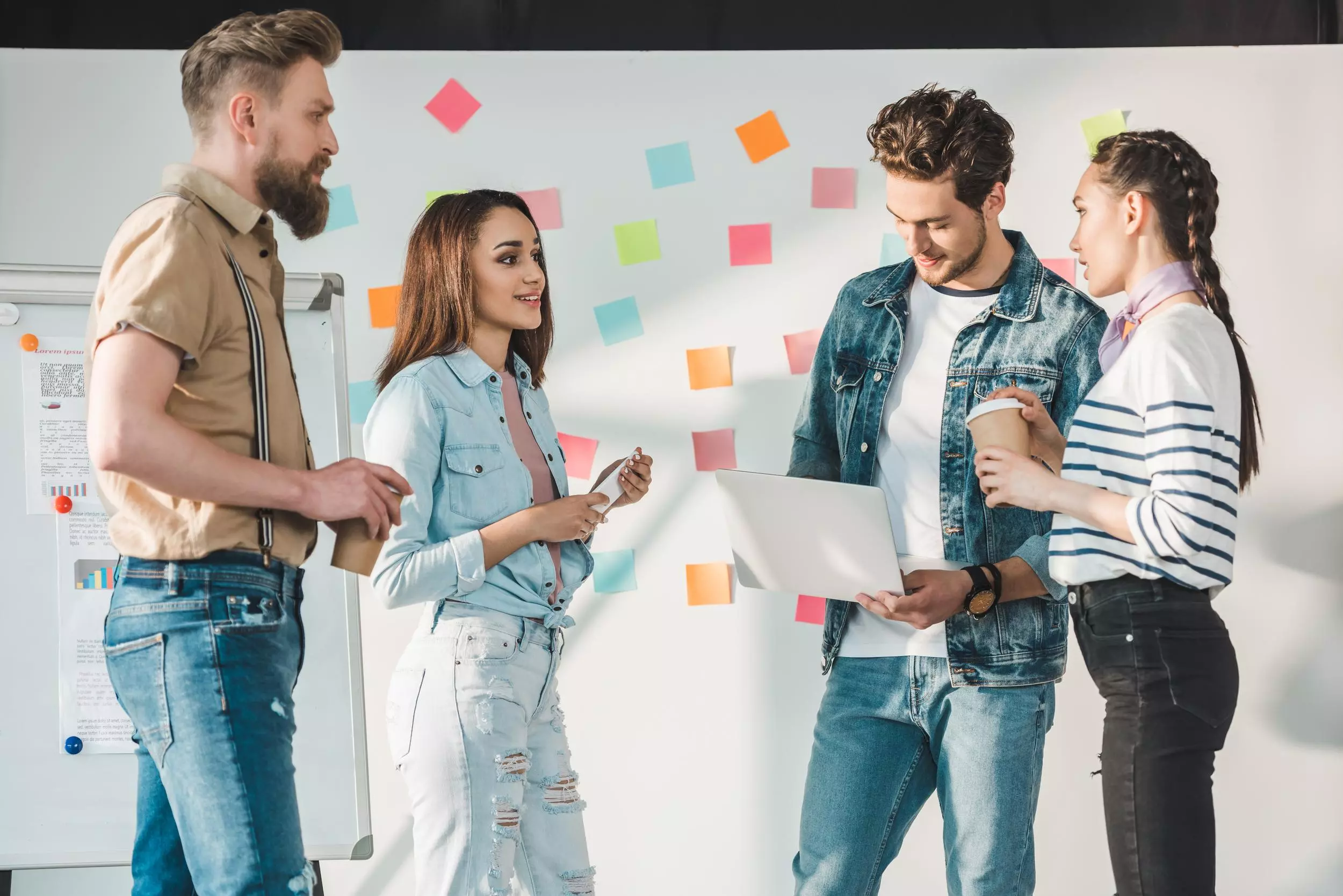 Diverse business team with laptop by white board with sticky notes in light workspace